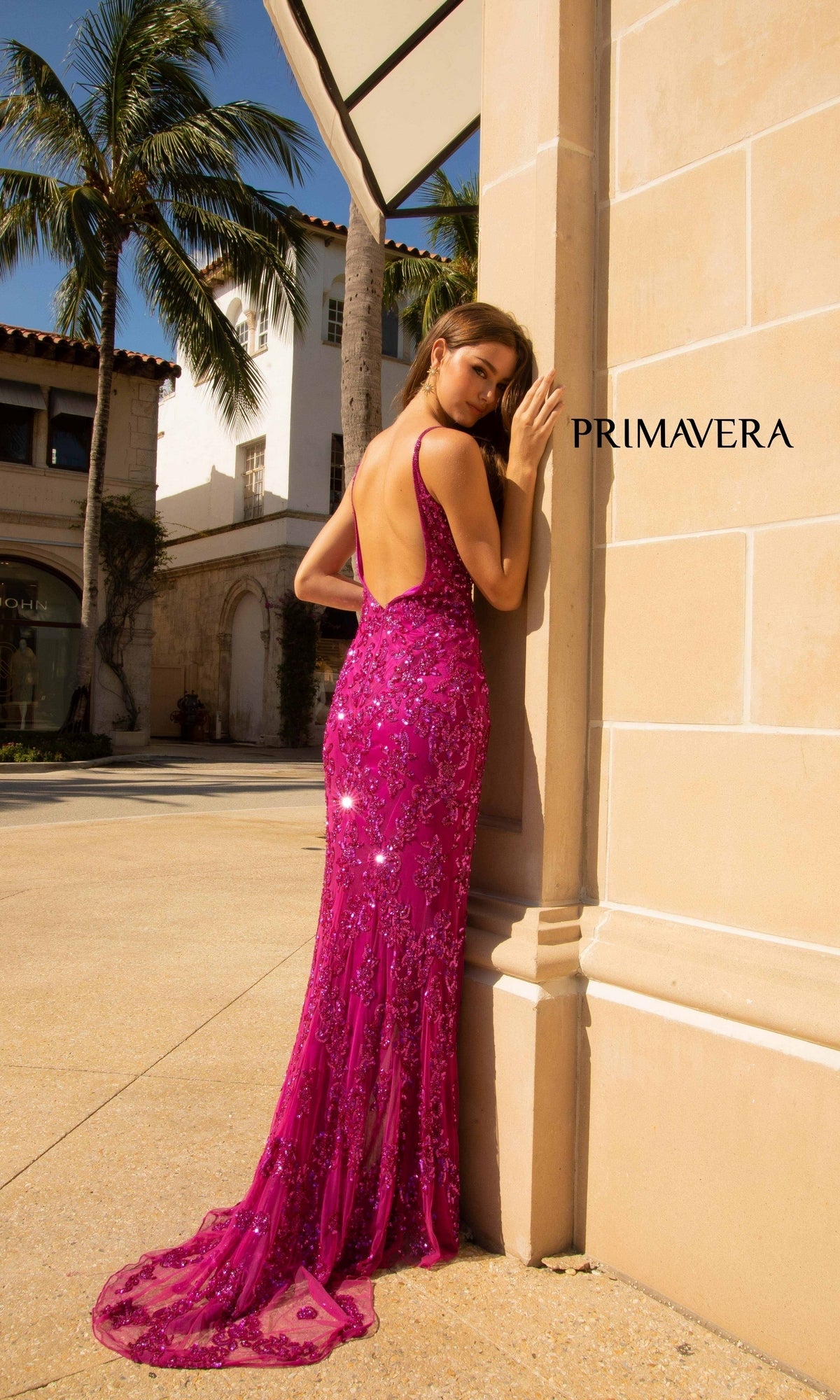 A woman in the Primavera 3913 Backless Long Sequin Prom Dress poses by a beige building under sunlight, her shimmering magenta gown catching every ray as